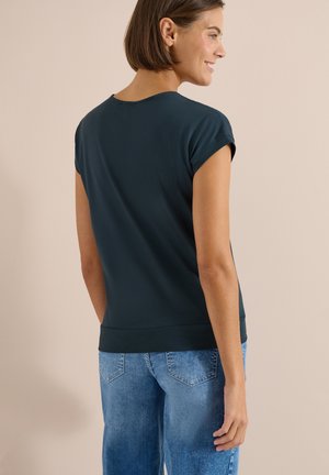 Femme aux cheveux courts et bruns, portant un haut vert foncé à manches courtes et un jean bleu, debout de profil devant un fond uni.