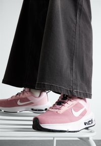 Sneakers Nike di colore rosa chiaro con tessuto in rete, dettagli neri e logo bianco. Indossati con pantaloni neri larghi. Ammortizzazione ad aria visibile.
