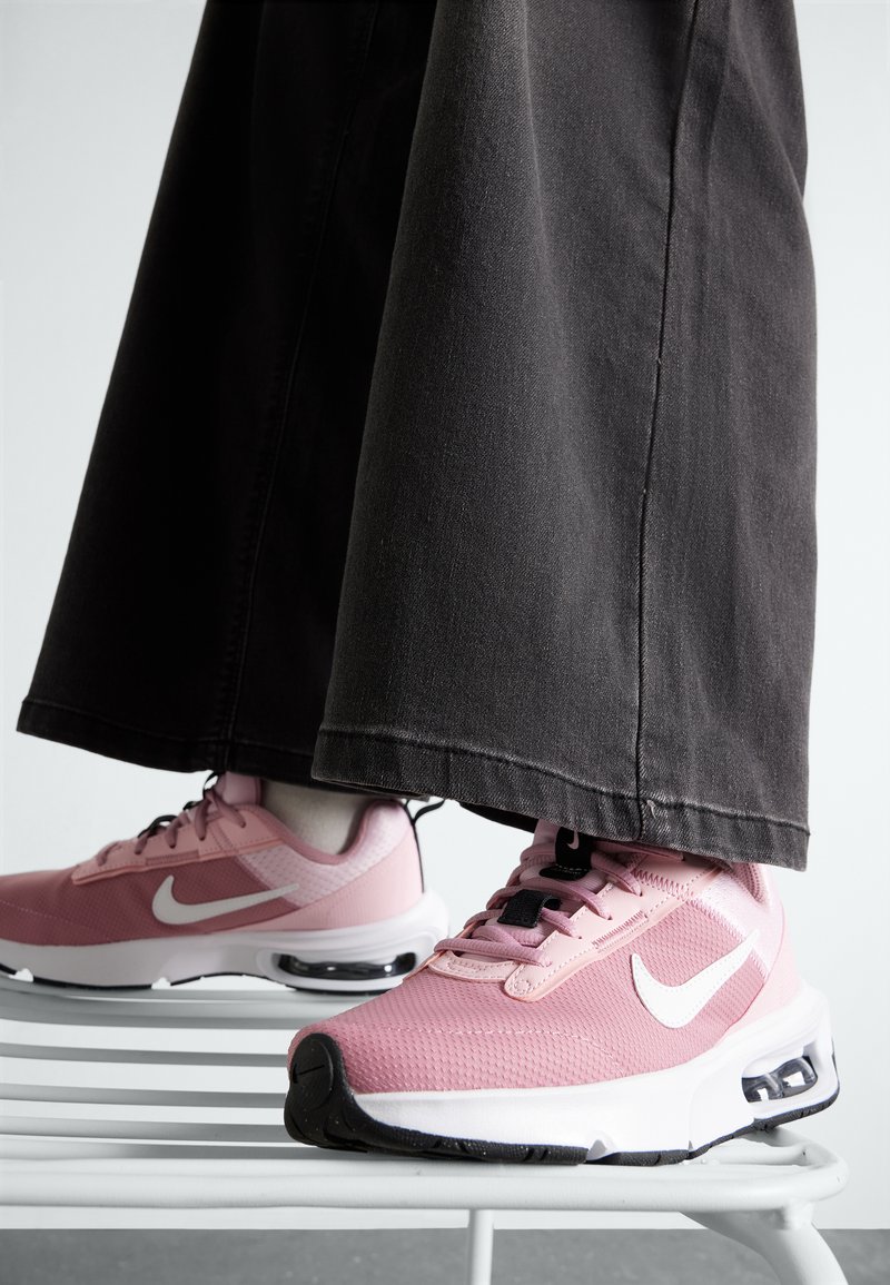 Sneakers Nike di colore rosa chiaro con tessuto in rete, dettagli neri e logo bianco. Indossati con pantaloni neri larghi. Ammortizzazione ad aria visibile.