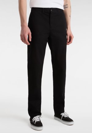 Homme portant un pantalon noir à jambes droites et des baskets basses noires avec des lacets blancs, debout devant un fond blanc.