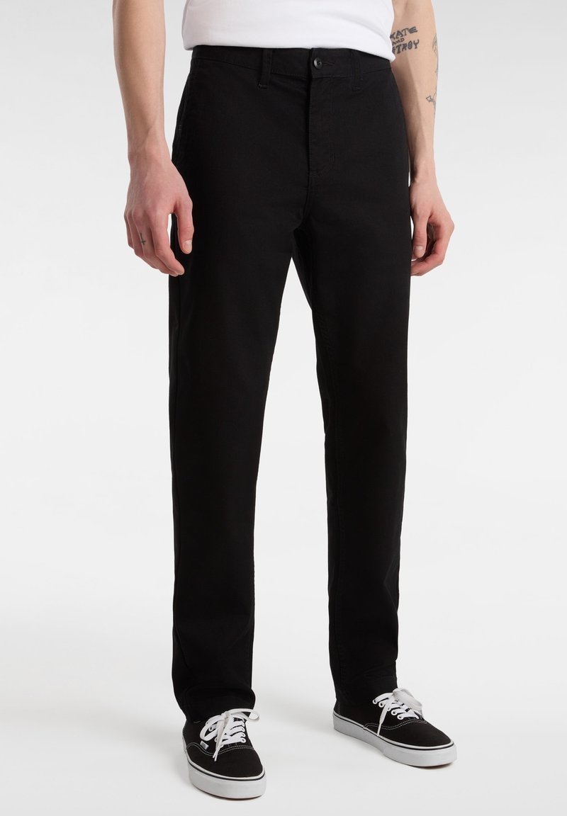 Homme portant un pantalon noir à jambes droites et des baskets basses noires avec des lacets blancs, debout devant un fond blanc.