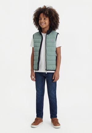 Χαμογελαστό παιδί με σγουρά μαλλιά που φοράει ανοιχτό πράσινο γιλέκο με βελούδινη υφή, λευκό μπλουζάκι, μπλε τζιν και καφέ αθλητικά παπούτσια, στέκεται μπροστά σε λευκό φόντο.