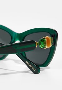 Ochelari de soare verzi, supradimensionați, cu lentile întunecate. Au un accent de cristal la balama și accesorii din metal auriu. Realizați dintr-un material plastic lucios.