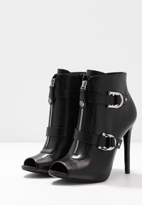 Czarne skórzane botki za kostkę z odkrytymi palcami, z metalowymi zamkami błyskawicznymi, akcentami klamer i wysokim obcasem w szpic. Gładka, błyszcząca faktura.