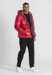 Rote Pufferjacke mit glänzender Oberfläche, schwarze Kapuzeninnenverkleidung mit Logotext, kombiniert mit schwarzen Hosen und weißen Sneakers.
