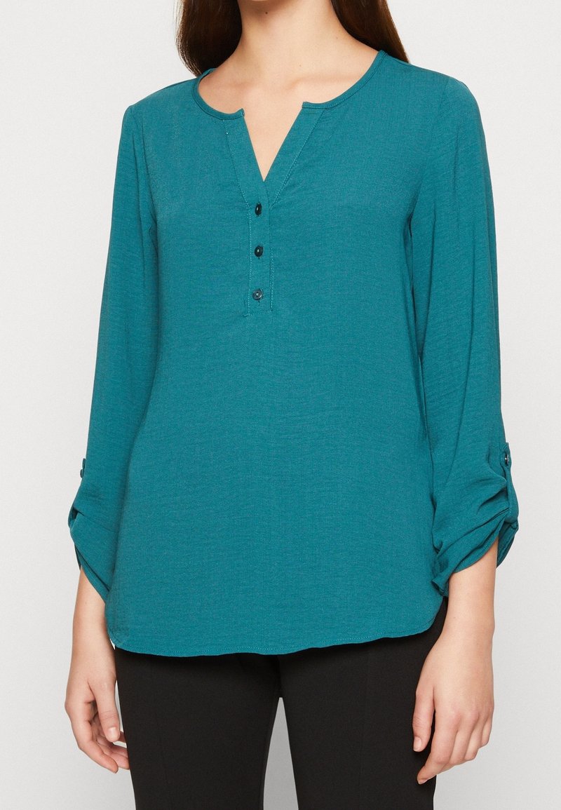 Femme portant un blouse en teal à manches longues avec un col en V boutonné et des manches retroussées, associée à un pantalon noir, se tenant devant un fond uni.