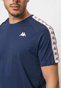 Námořnické modré tričko Kappa s krátkými rukávy, které má bílý pruh na ramenou zdobený oranžovými logy a malé bílé logo na hrudi.