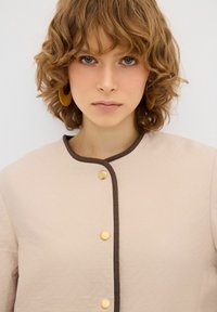 Giacca trapuntata beige con rifiniture marroni e bottoni dorati, caratterizzata da un collo rotondo e un design in tessuto testurizzato.