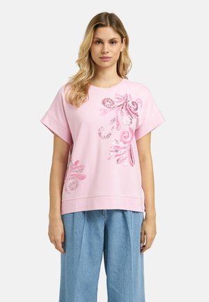 Woman wearing light pink short-sleeve top with pink floral sequin design, paired with loose-fitting light blue jeans, standing against white background.