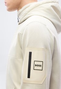 Personne portant une veste à capuche beige clair avec une manche texturée et une poche plaquée dotée d'une fermeture éclair verticale et du mot BOSS.