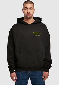 Hanorac negru din țesătură groasă, cu o pockets frontal. Are text verde aprins și o glugă pentru căldură. Design simplu și croială lejeră.