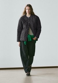 Kvinde, der går iført en sort quiltet jakke, grå skjorte, grøn sweater bundet om livet, løse sorte bukser og mørke sko foran en ensfarvet baggrund.