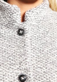 Grijze en witte textuurjack met een hoge kraag, voorzien van zwarte knopen en een geweven patroon. Zichtbare stikdetails doorheen.