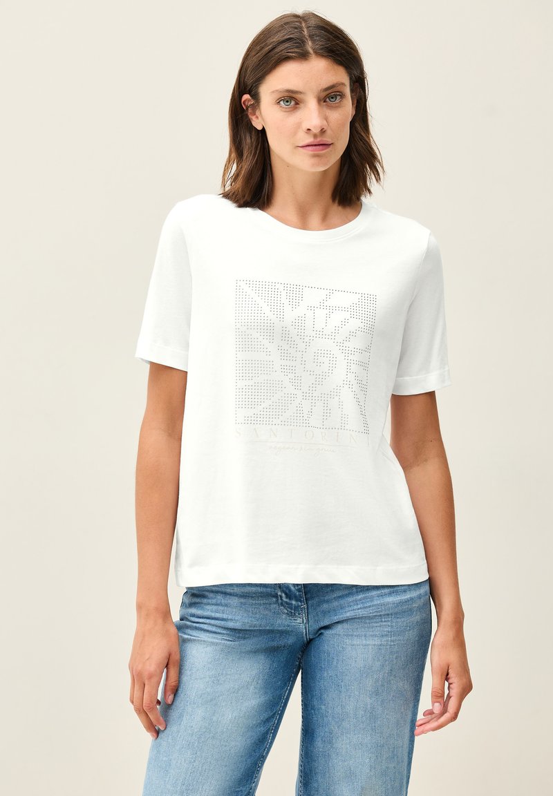 T-shirt bianco in cotone con collo rotondo, maniche corte e un motivo geometrico a pois sul davanti. Abbinato a jeans di denim blu.