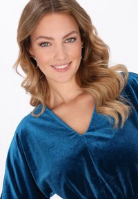 Türkiz bársony ruha V-nyakkivágással és bő ujjakkal. Lágy tapintású és finom fényű anyagból készült. A modell merész fekete fülbevalót visel.