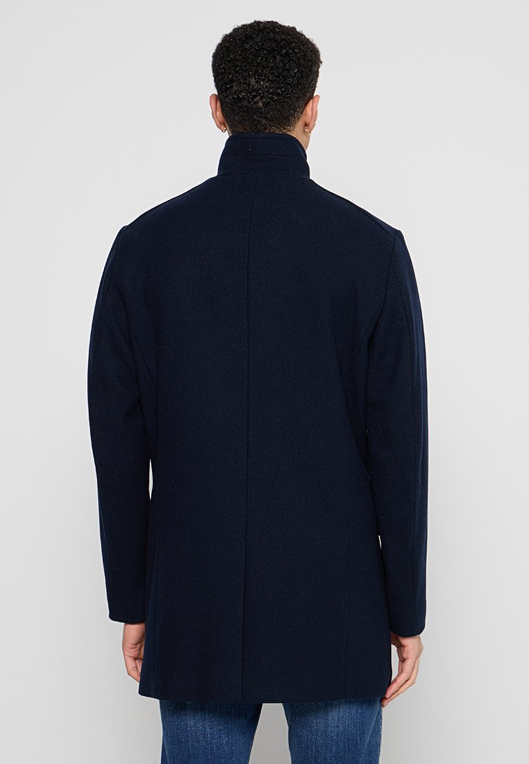 Marineblauw wollen blend jas met een hoge kraag, een getailleerde pasvorm en een gestructureerd visgraatpatroon, met een nette achterkant en zonder zichtbare hardware.