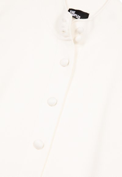 Gros plan sur un vêtement blanc avec des boutons recouverts de tissu le long de la patte de boutonnage et une étiquette de col portant la mention "THE PEOPLES".
