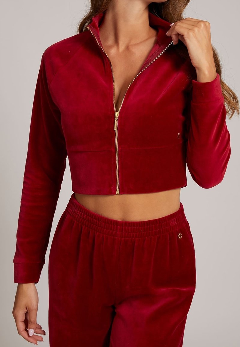 Femme portant une veste courte zippée en velours rouge et un pantalon assorti à taille élastique, laissant entrevoir le ventre et tenant le col de la veste.