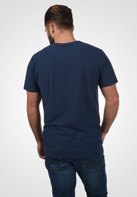 Tamno plava majica s kratkim rukavima, izrađena od pamuka, s okruglim izrezom i opuštenim krojem, nosi se s plavim trapericama, prikazana sa stražnje strane.