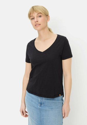 Blonde vrouw met een zwart V-hals shirt met korte mouwen en een lichtblauwe spijkerrok, staand tegen een witte achtergrond.