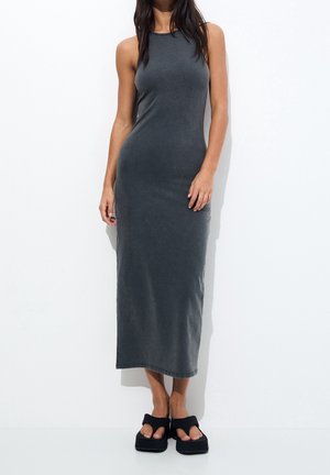 Woman wearing a sleeveless, fitted gray maxi dress and black platform sandals standing against a white background.