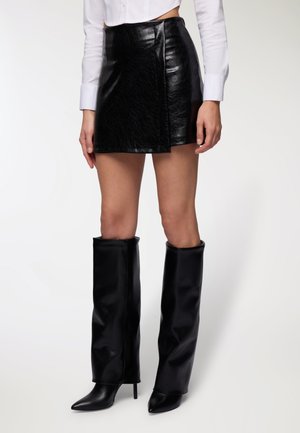 Mini gonna in pelle nera con una finitura testurizzata, caratterizzata da un design con sovrapposizione frontale. Abbinata a stivali neri alti e con tacco a punta.