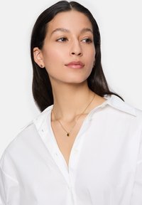 Goldschmuck mit runden Ohrsteckern und einer zarten Halskette mit einem ovalen Anhänger, getragen zu einem weißen Hemd mit Knöpfen.