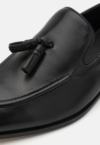 Mocasines de cuero negro con una textura suave, punta redondeada y adornos de borlas decorativas. Costuras detalladas a lo largo de los bordes.