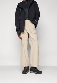 Black waterproof jacket, beige wide-leg pants with drawstring, and black hiking boots with textured surface and laces.