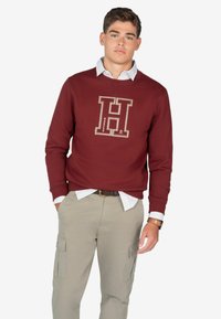 Bordowy sweatshirt z dużym haftowanym logo "H" i kontrastowymi szwami, w połączeniu z koszulą z kołnierzykiem i beżowymi spodniami cargo.
