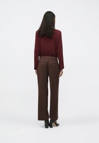 Burgundi hosszú ujjú felsőt, barna széles lábú nadrágot és fekete tömbsarkú cipőt viselő göndör hajú nő egy fehér háttér előtt áll.