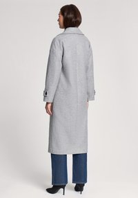 Szary wełniany płaszcz z tylnym rozcięciem, opadającymi ramionami i dużymi guzikami przy mankietach. Połączony z szerokimi spodniami dżinsowymi i czarnymi obcasami.