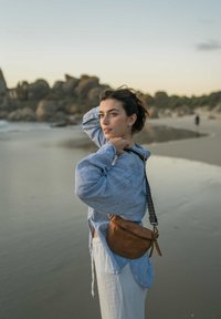 Junge Frau mit brauner Ledertasche, die sich die Haare zurechtmacht, steht am Sandstrand mit Felsen im Hintergrund bei Sonnenuntergang.