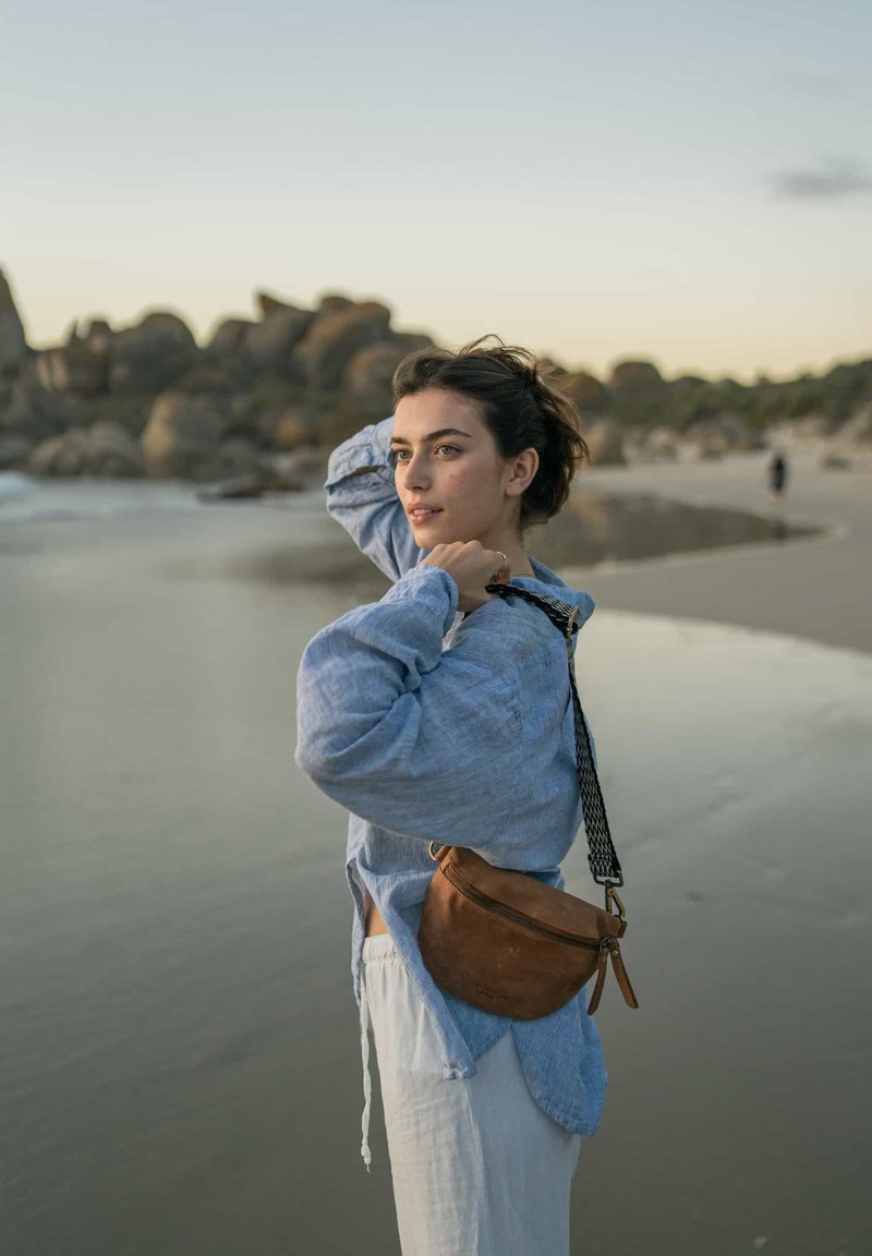 Junge Frau mit brauner Ledertasche, die sich die Haare zurechtmacht, steht am Sandstrand mit Felsen im Hintergrund bei Sonnenuntergang.