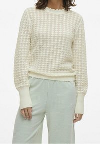 Pull en maille couleur crème avec un motif de diamants texturé, des poignets côtelés et un col festonné, assorti à un pantalon à carreaux vert clair.