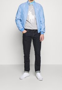 Ljusblå randig bomberjacka med stickade ärmslut, grå sweatshirt med ett vitt grafiskt tryck, mörk denim jeans och vita sneakers med grå detaljer.