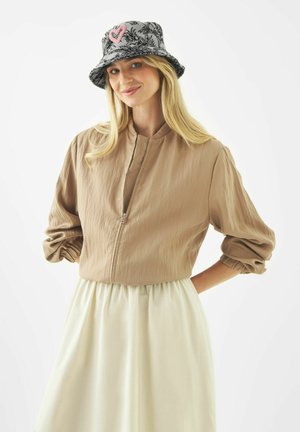 Femme souriante portant une veste beige zippée, une jupe blanche et un chapeau seau noir à motif avec un détail de cœur rose, debout devant un fond blanc.