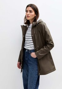 Impermeabile color verde oliva con cappuccio, chiusura con zip e corde regolabili, abbinato a una camicia a righe bianche e navy e jeans blu.