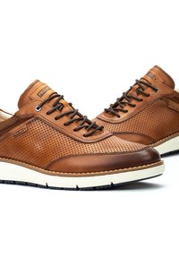 Zapatillas de cuero marrón con diseño perforado, punta redondeada, cordones oscuros y suelas de color claro. Presenta una marca en el lateral.