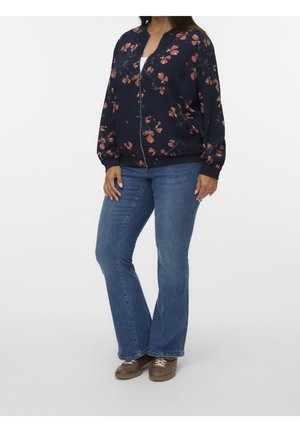 Femme portant une veste zippée bleu marine à motif floral, un jean évasé bleu et des baskets marron, debout devant un fond uni.