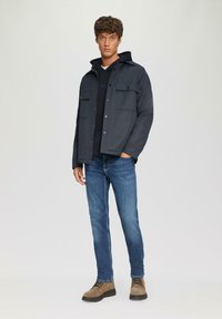 Marinejas met drukknoopjes en voorkruiemen, gelaagd over een zwarte hoodie. Gedragen met blauwe jeans en tan veterschoenen.