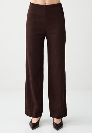 Femme portant un pantalon large taille haute marron foncé associé à des chaussures pointues foncées, debout devant un fond clair uni.