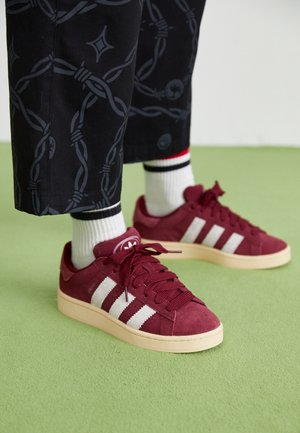 Osoba nosząca czarne spodnie z wzorem z drutu kolczastego, białe skarpetki z czarnymi i czerwonymi paskami oraz bordowe buty sportowe Adidas na zielonej podłodze.