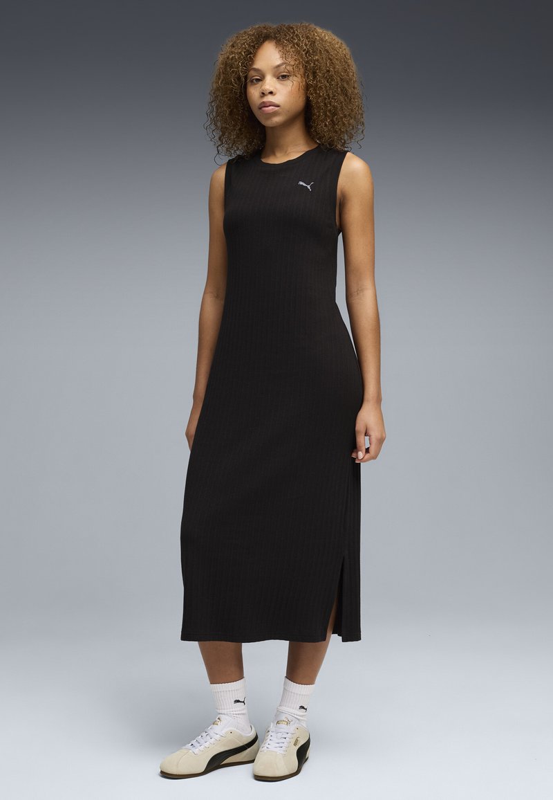 Femme aux cheveux bouclés portant une robe midi noire sans manches avec une fente sur le côté, des chaussettes blanches Puma et des baskets beige Puma, debout sur un fond gris.