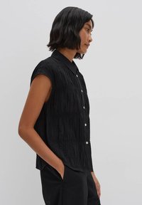 Maglietta nera senza maniche con colletto, caratterizzata da un design testurizzato e plissettato e bottoni argentati lungo il davanti.