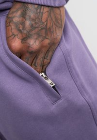Een close-up van een paarse kledingstuk met een ritszak. Het heeft een gestructureerde stof en zilveren hardware, met een zichtbare getatoeëerde hand die de zak vasthoudt.