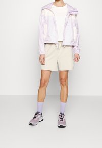 Lichtpaarse zip-up jacket met beige accenten, gecombineerd met beige shorts, paarse sokken en multikleurige sportschoenen. Gladde textuur, casual ontwerp.