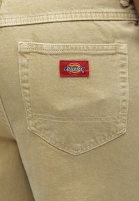 Tissu beige avec une poche arrière distincte ornée d'un écusson au logo Dickies rouge et jaune. La poche a une forme simple et angulaire avec des bords cousus.