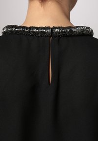 Zwarte kledingstuk met een bewerkte, textuur-kragen en een sleutelgatopening aan de achterkant. Gladde stof met een matte afwerking.