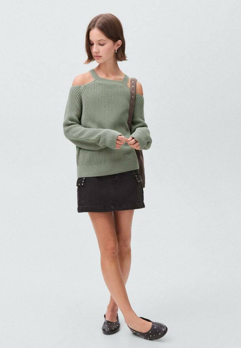 Grüner gestrickter Pullover mit Schulterausschnitten, schwarzer Minirock mit Taschen und schwarze Slipper mit silbernen Nieten.